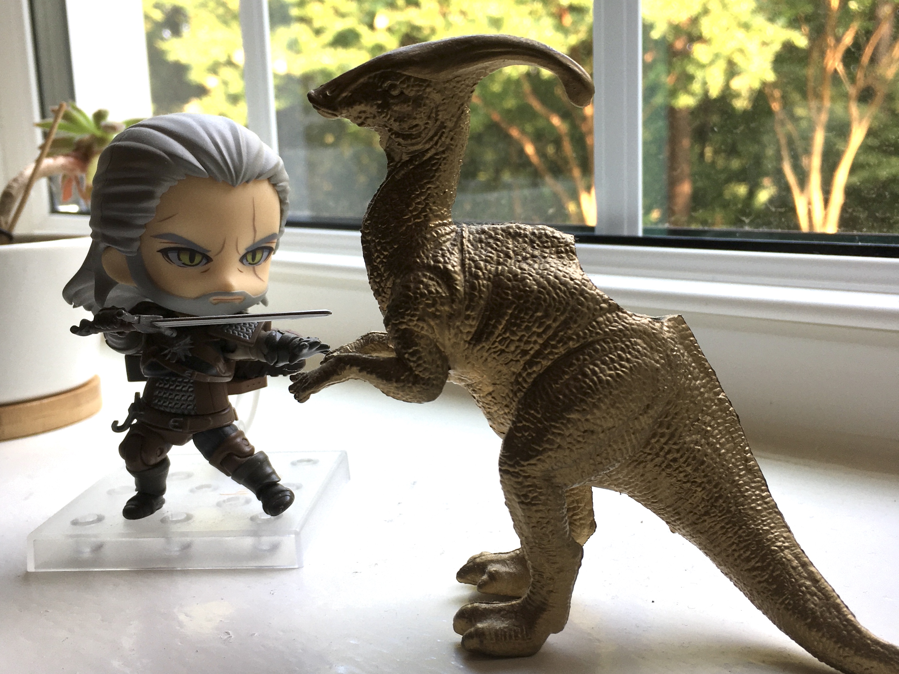 A chibi Geralt figure holding a sword and a gold plastic dinosaur planter on a windowsill, facing each other as if in battle