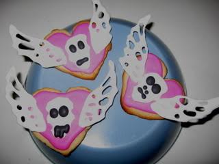 Three heart-shaped cookies on an overturned bowl. The cookies are topped with rudimentary sugar skull and wing decorations.